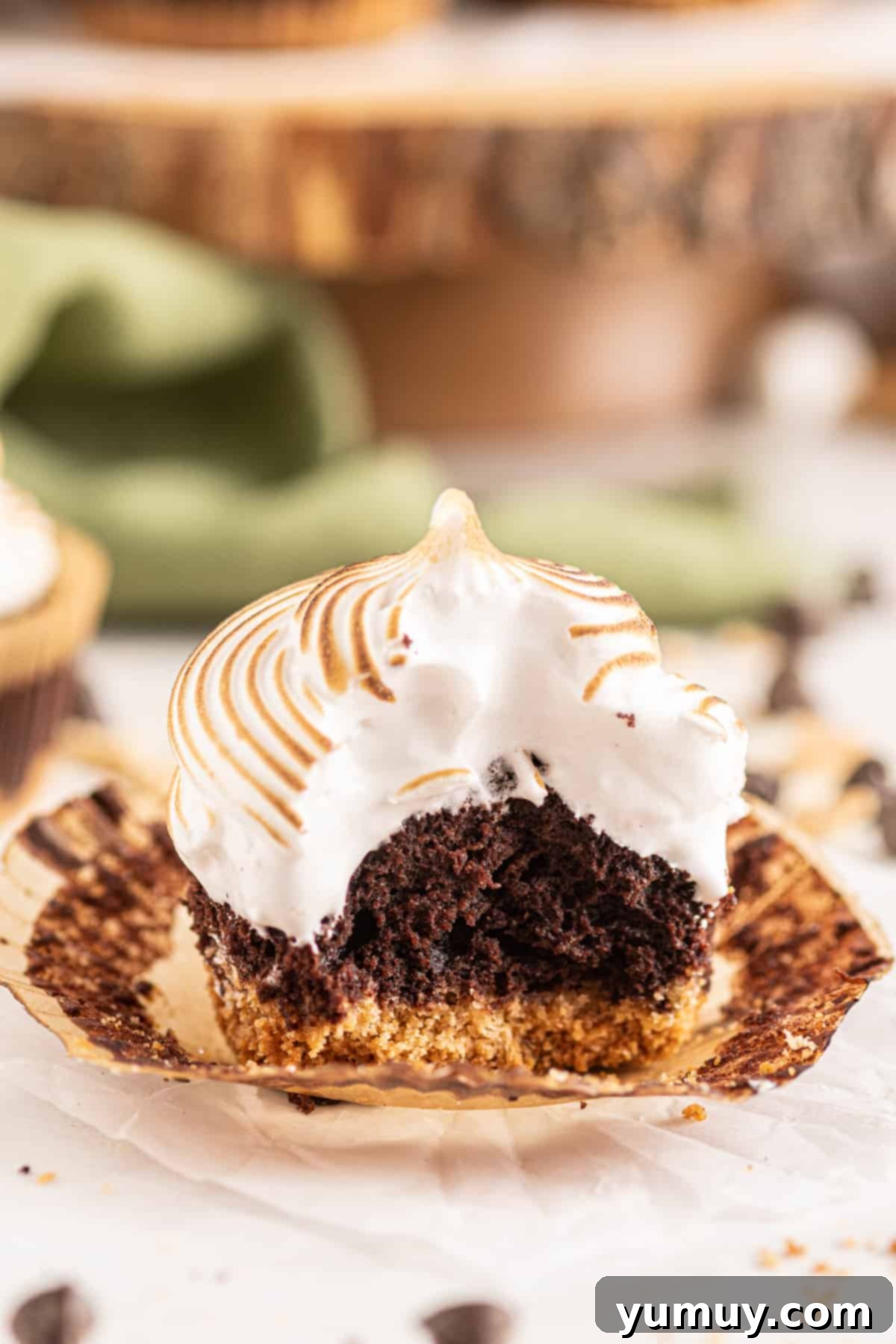 Close up side view of an unwrapped and bitten s'mores cupcake, revealing its chocolate cake, graham cracker crust, and fluffy marshmallow meringue topping.