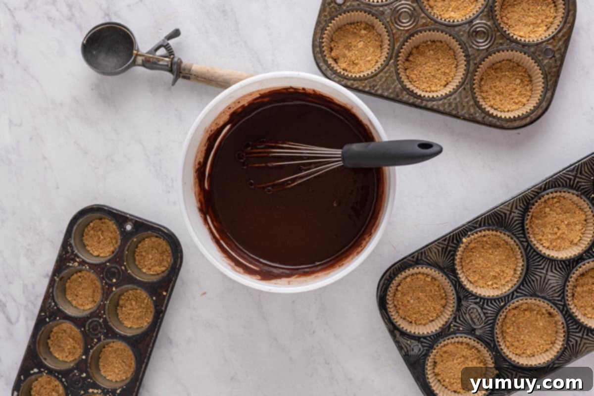 Chocolate cupcake batter in a white mixing bowl with a whisk, surrounded by cupcake tins filled with graham cracker crust.