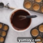 Chocolate cupcake batter in a white mixing bowl with a whisk, surrounded by cupcake tins filled with graham cracker crust.