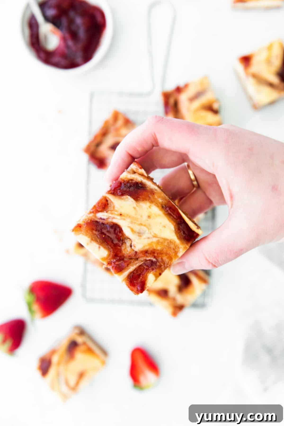 Luscious Strawberry Cheesecake Bars 2 Overhead view of a hand holding a strawberry cheesecake bar, showcasing its creamy texture and strawberry swirl.