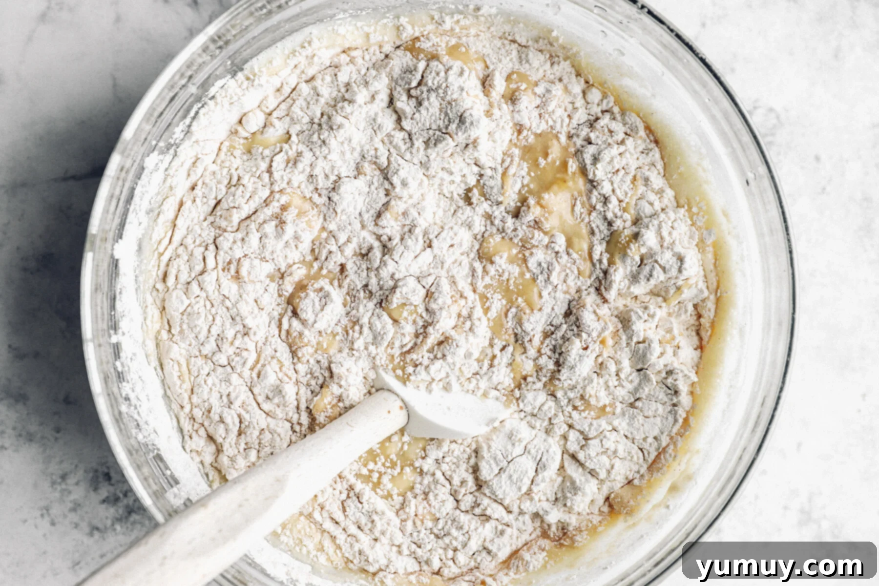 dry ingredients being folded into wet ingredients in a glass bowl with a rubber spatula.