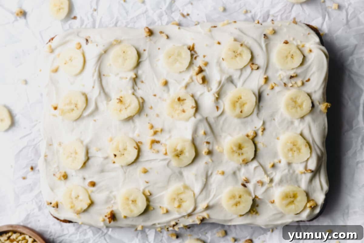 overhead view of banana cake on a white table with banana slices and crushed nuts on top.