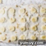 overhead view of banana cake on a white table with banana slices and crushed nuts on top.