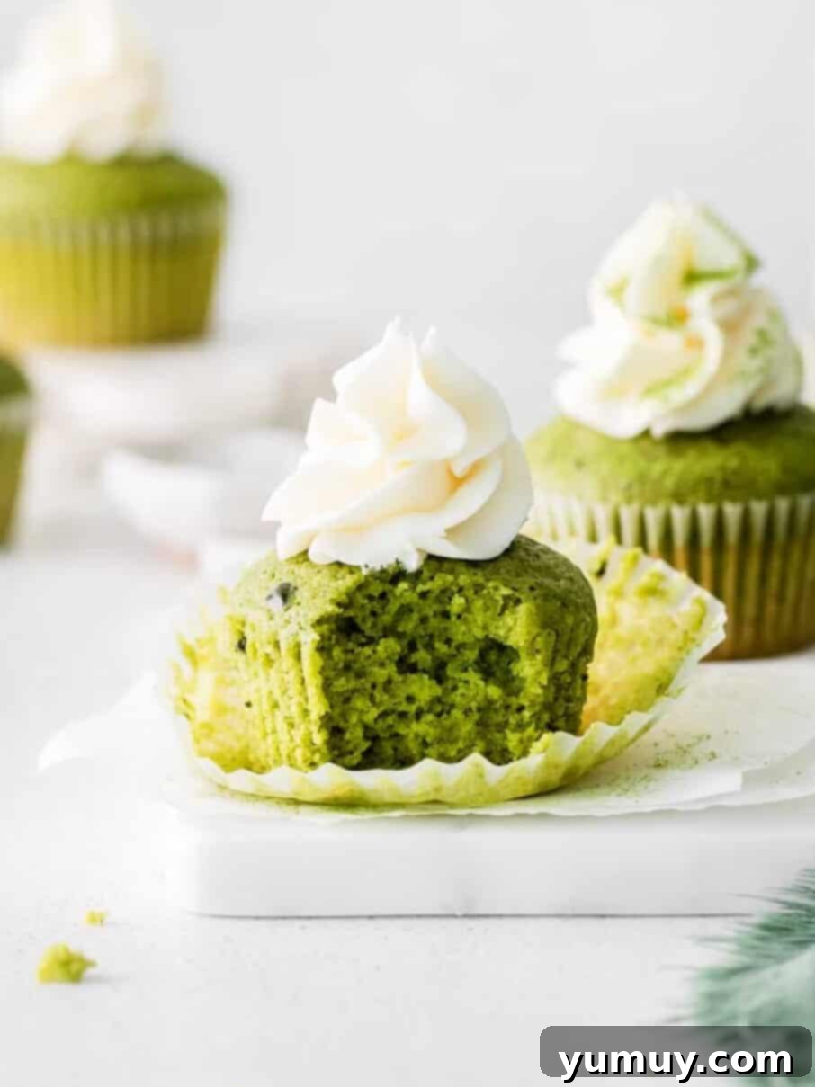 Two vibrant green matcha cupcakes with creamy white frosting, one with a bite taken, resting on parchment paper on a marble tray.