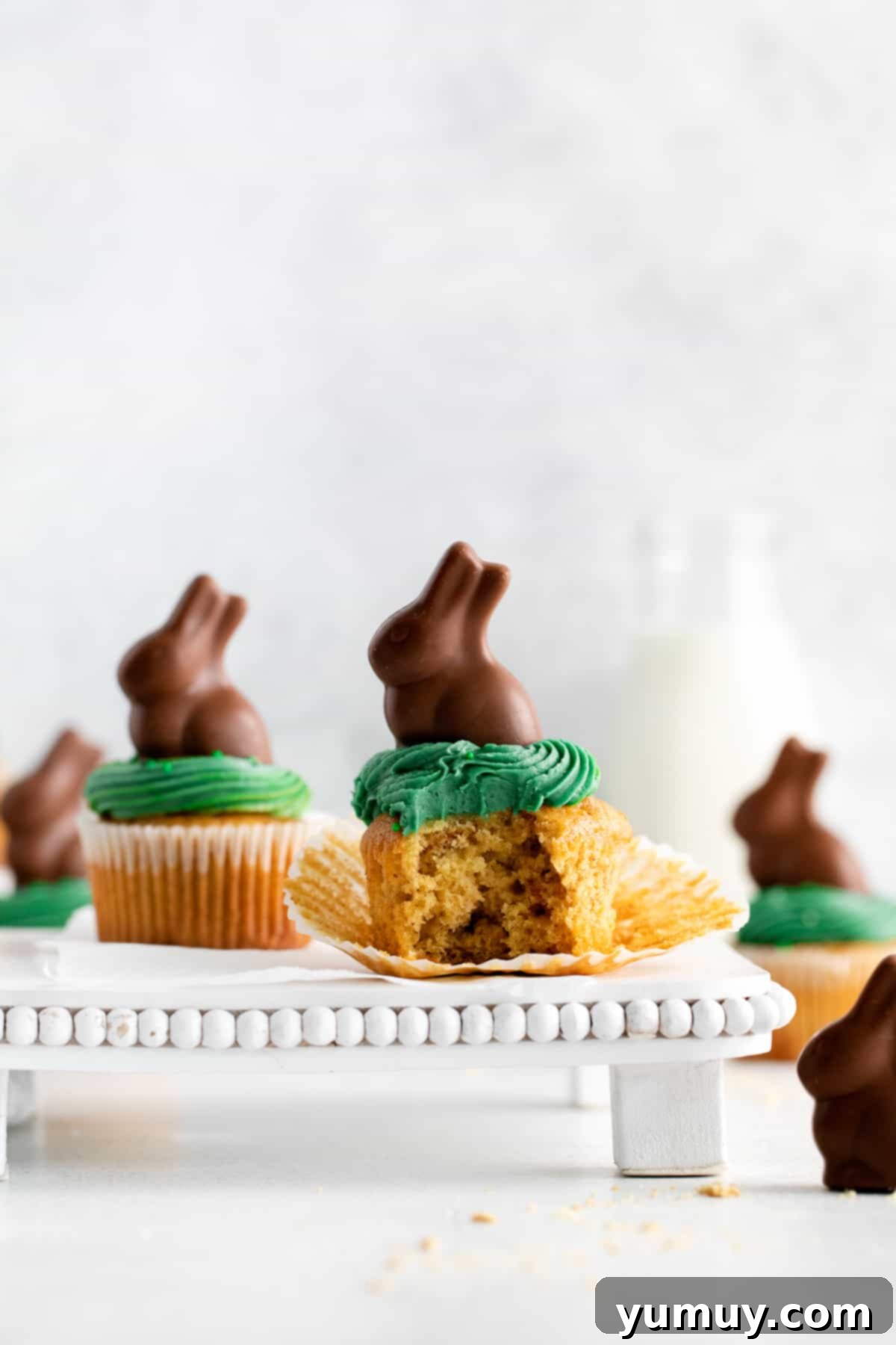 Two beautifully decorated Easter bunny cupcakes on a pristine white cake stand, one showing a perfect bite.