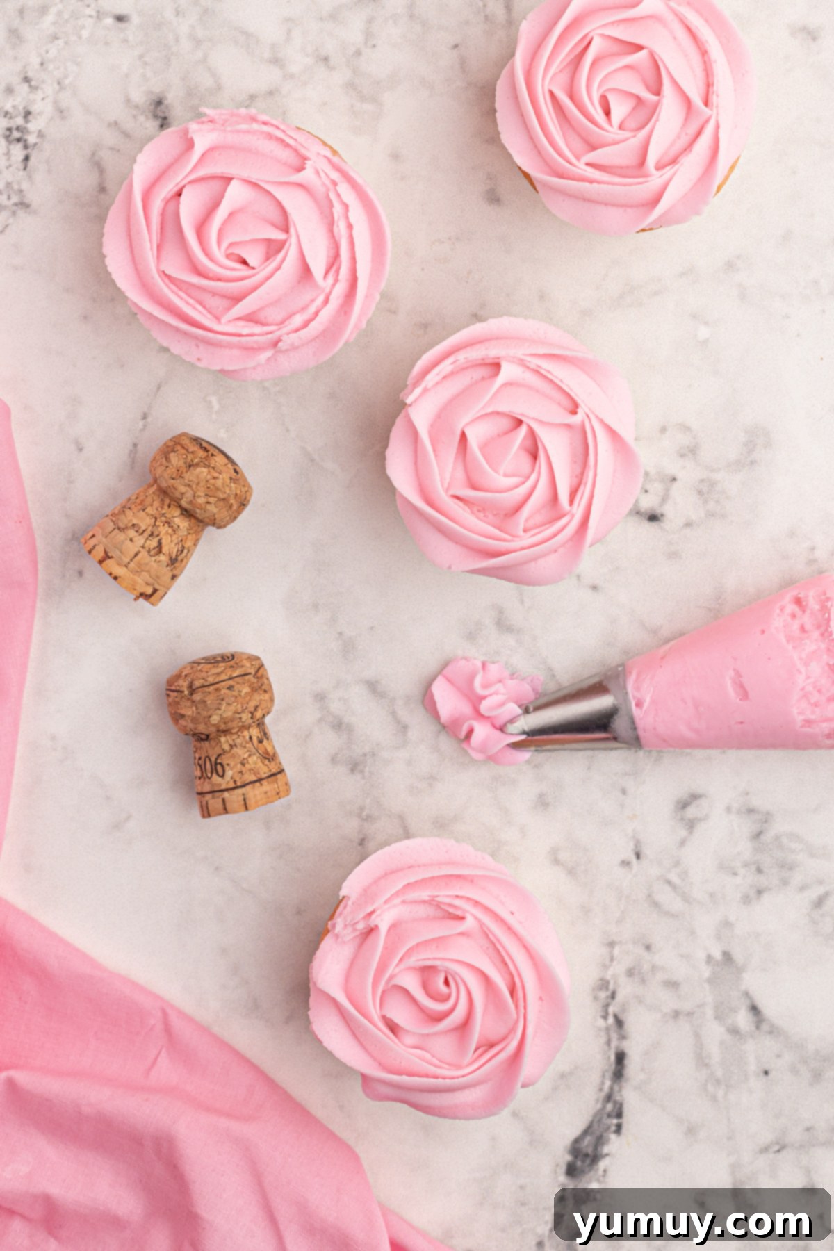 Overhead view of four pink moscato cupcakes, perfectly frosted, alongside a piping bag and champagne corks, ready for a celebration.