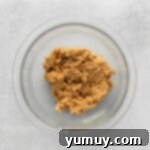 Peanut butter in a glass bowl on a gray background, showing chopped peanuts being folded in.