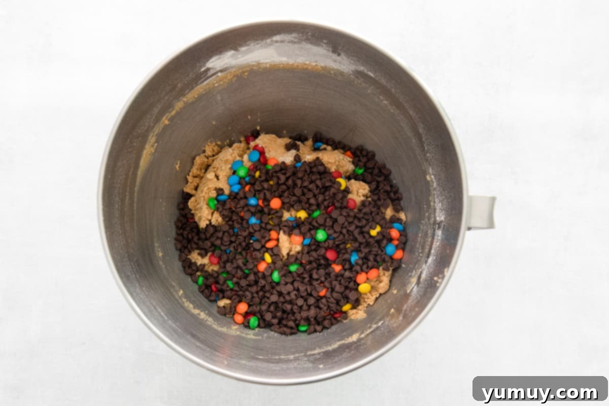 No-Bake Monster Cookie Dough 9 A metal bowl brimming with a colorful assortment of chocolate chips and M&Ms, ready to be folded into the dough.