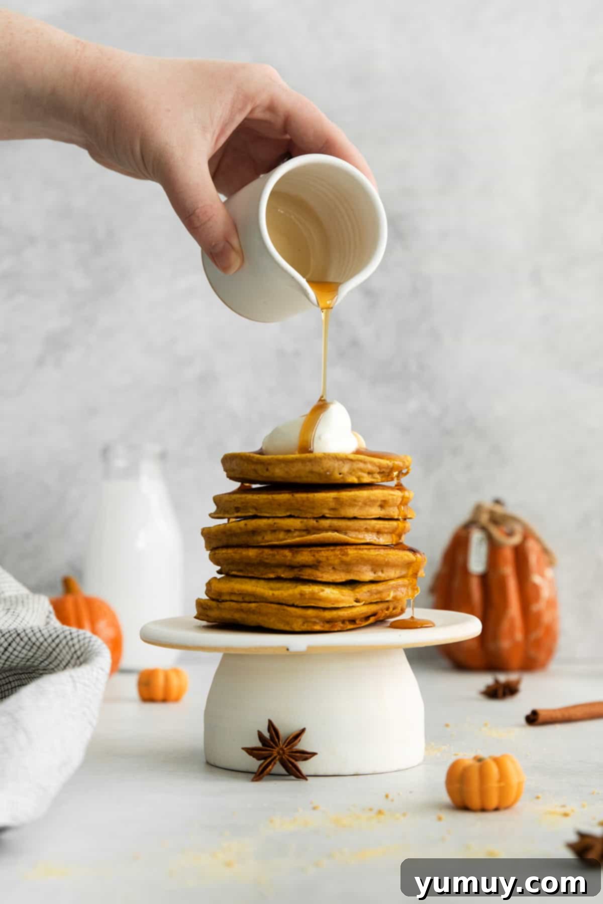 A person pouring maple syrup generously over a tall stack of golden pumpkin pancakes, ready to be enjoyed.