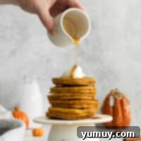 A person pouring a steady stream of golden maple syrup onto a stack of warm pumpkin pancakes.