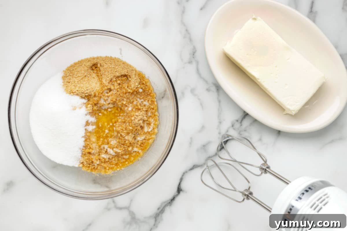 A bowl with graham cracker crumbs, melted butter, and granulated sugar, next to a hand mixer, ready for mixing the crust.