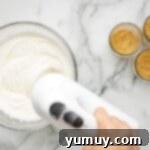 A hand mixer blending cream cheese, powdered sugar, and vanilla extract in a bowl until smooth.