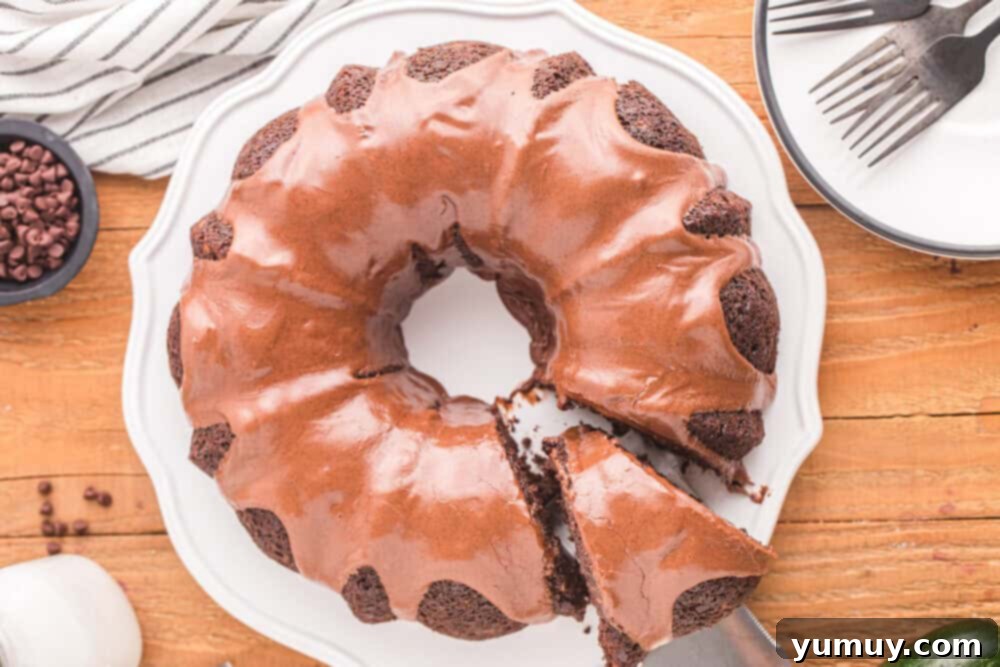 Luscious Chocolate Zucchini Bundt 3 Overhead view of a glazed chocolate zucchini bundt cake on a white cake stand with a slice being removed by a cake server.