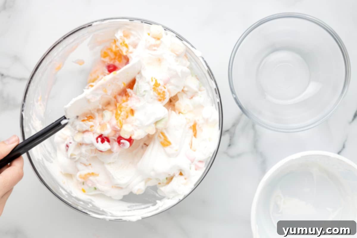 Ambrosia salad being gently mixed with a rubber spatula in a glass bowl, showcasing its fluffy texture.