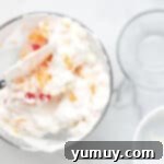 Ambrosia salad being gently mixed with a rubber spatula in a glass bowl.