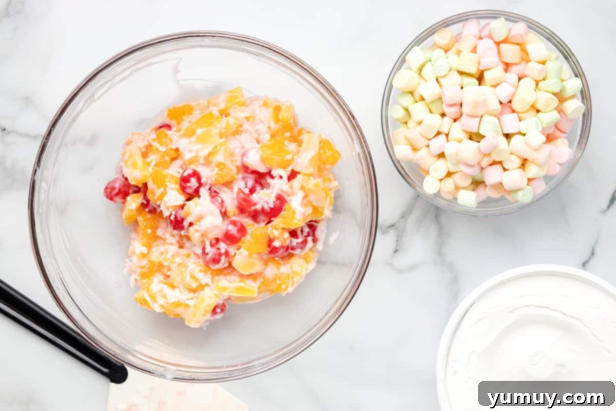Mixed canned fruits in a glass bowl with vanilla yogurt and coconut flakes, ready for the next step of ambrosia preparation.