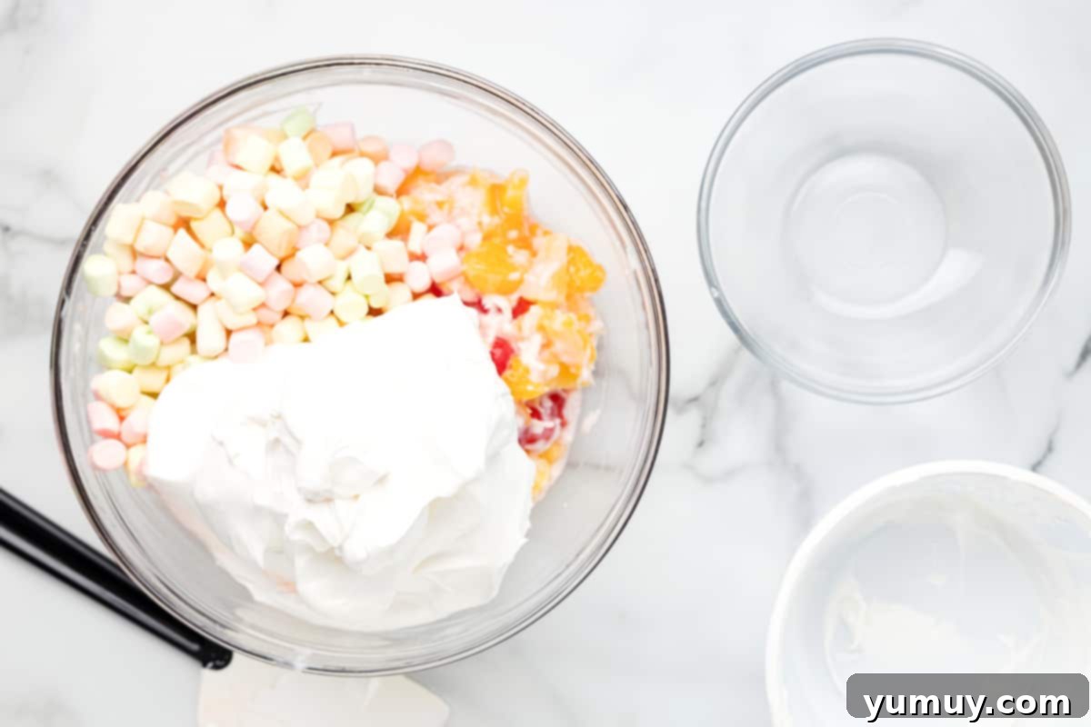 Cool Whip and mini marshmallows being gently folded into the fruit and yogurt mixture in a glass bowl for ambrosia salad.