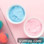 2 bowls of frosting for cotton candy cupcakes in white bowls, one colored blue and one colored pink.