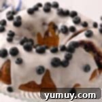 Bursting Blueberry Bundt 15 Removing a slice from a glazed blueberry bundt cake topped with fresh blueberries on a glass cake stand.