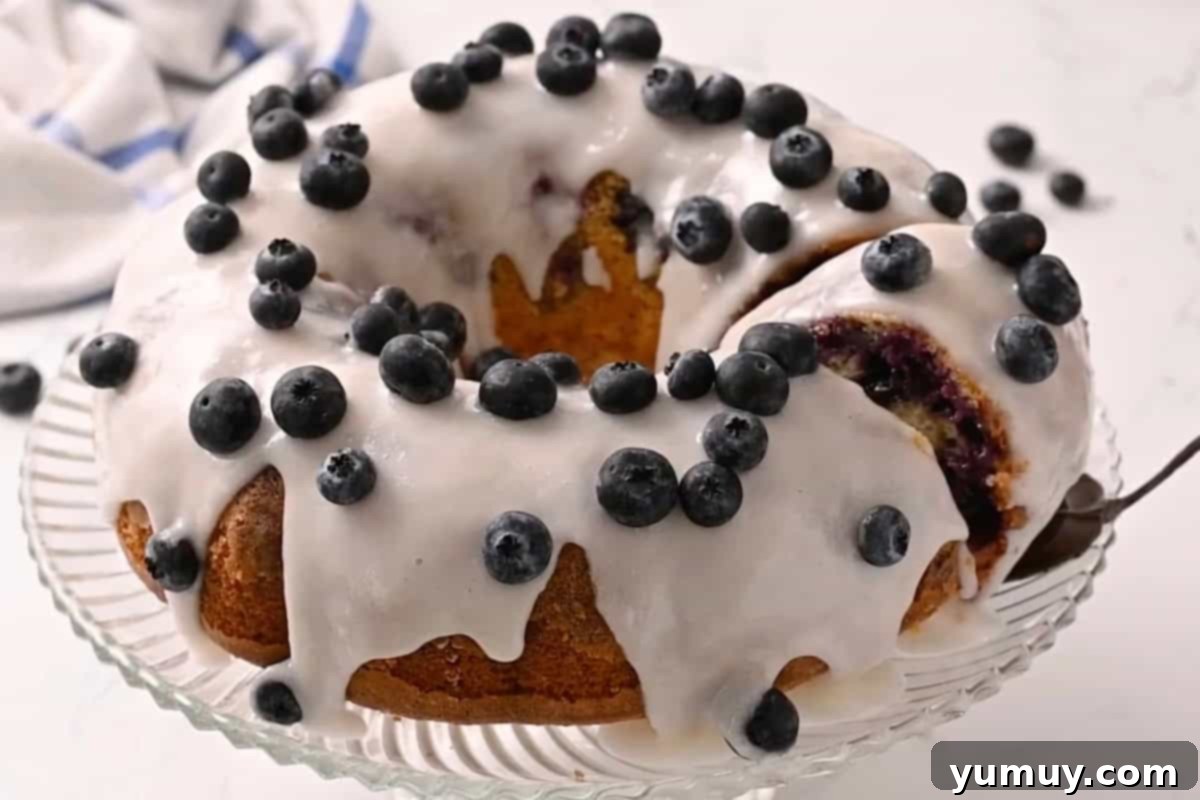 Bursting Blueberry Bundt 22 A slice being carefully removed from a glazed blueberry bundt cake, adorned with fresh blueberries on a glass cake stand.