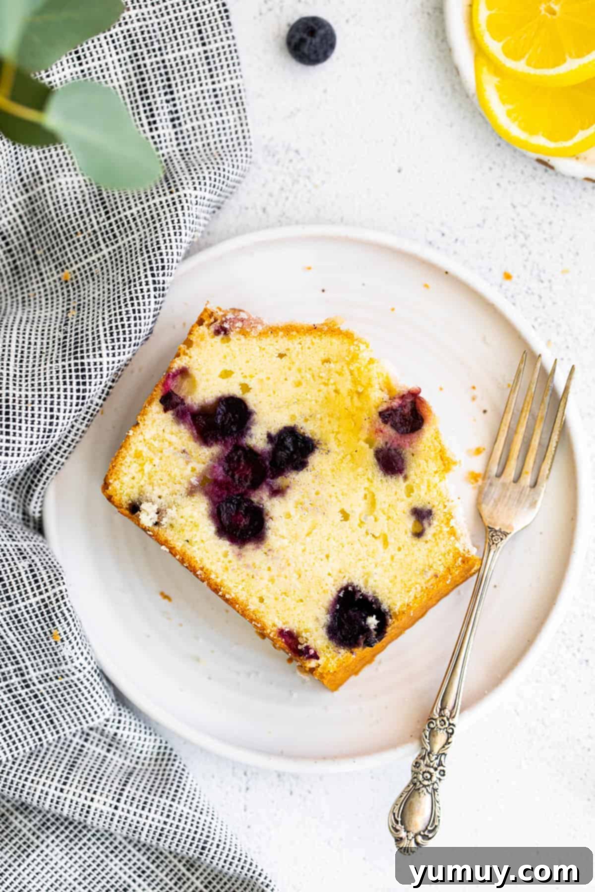 A close-up view of a single slice of lemon blueberry pound cake on a white plate, highlighting the vibrant blueberries and moist texture.