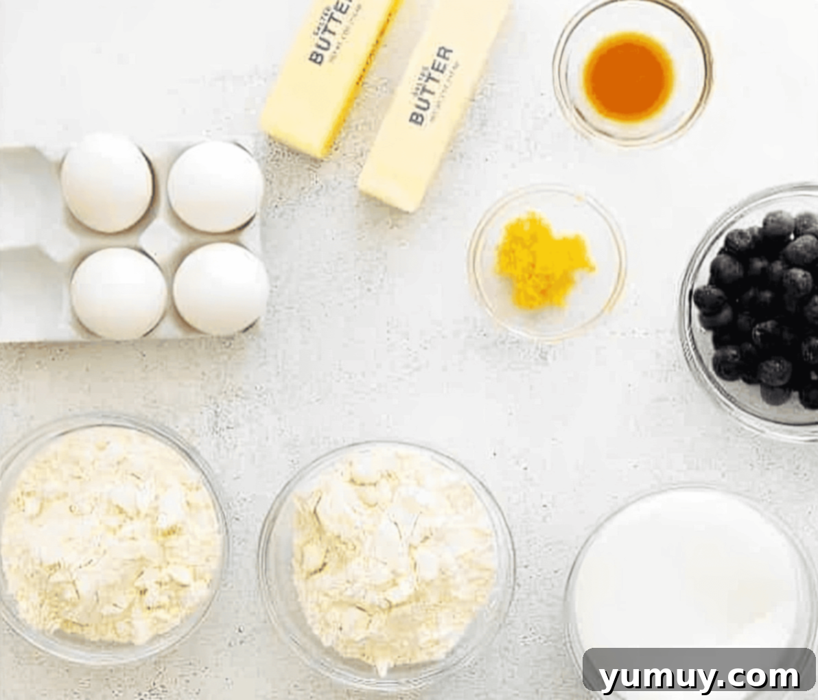 A beautiful flat lay of all ingredients required for making lemon blueberry pound cake, neatly arranged on a white surface.
