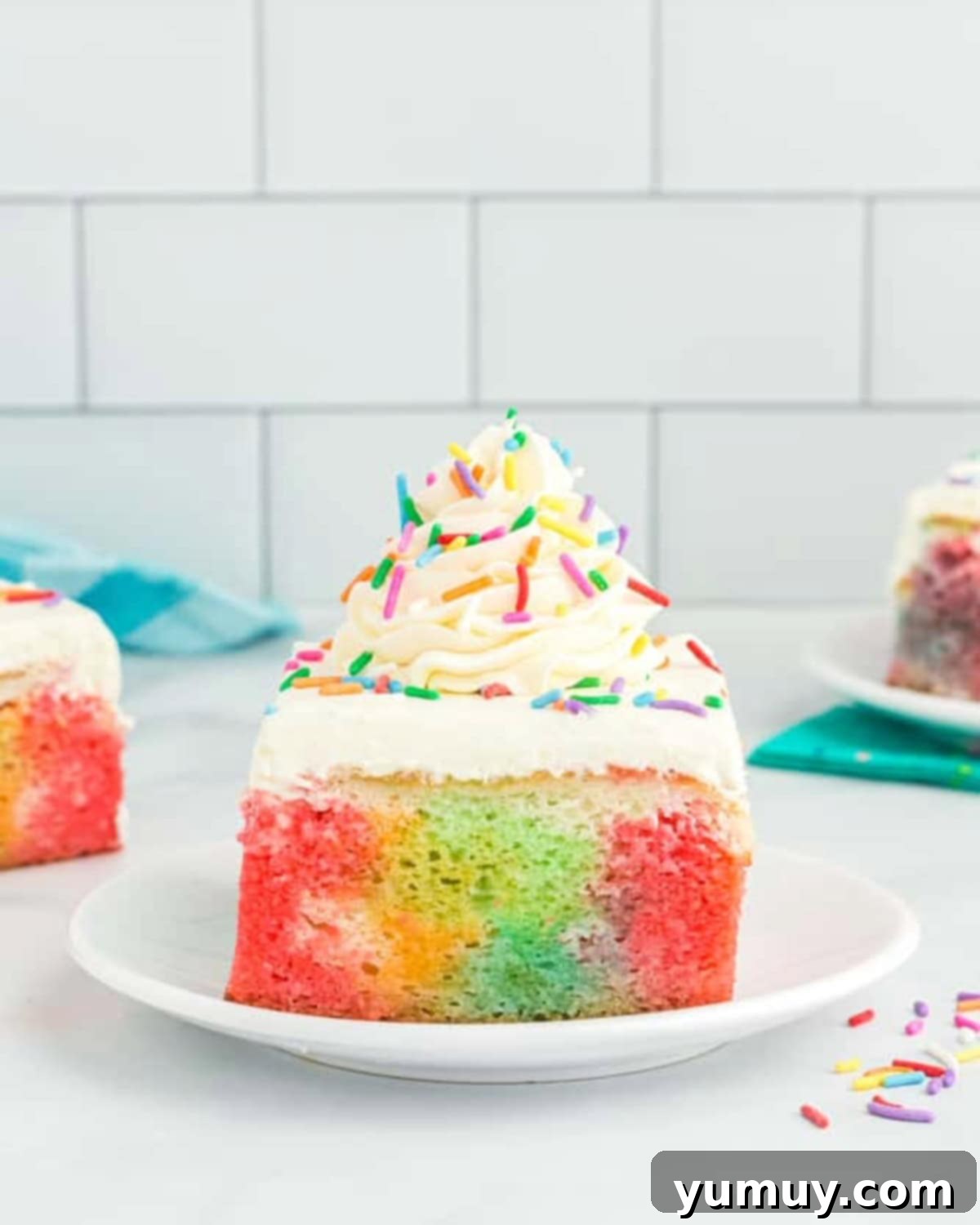 A vibrant slice of rainbow Jell-O poke cake on a white plate, revealing layers of colorful gelatin soaked into the vanilla cake, topped with creamy white frosting.