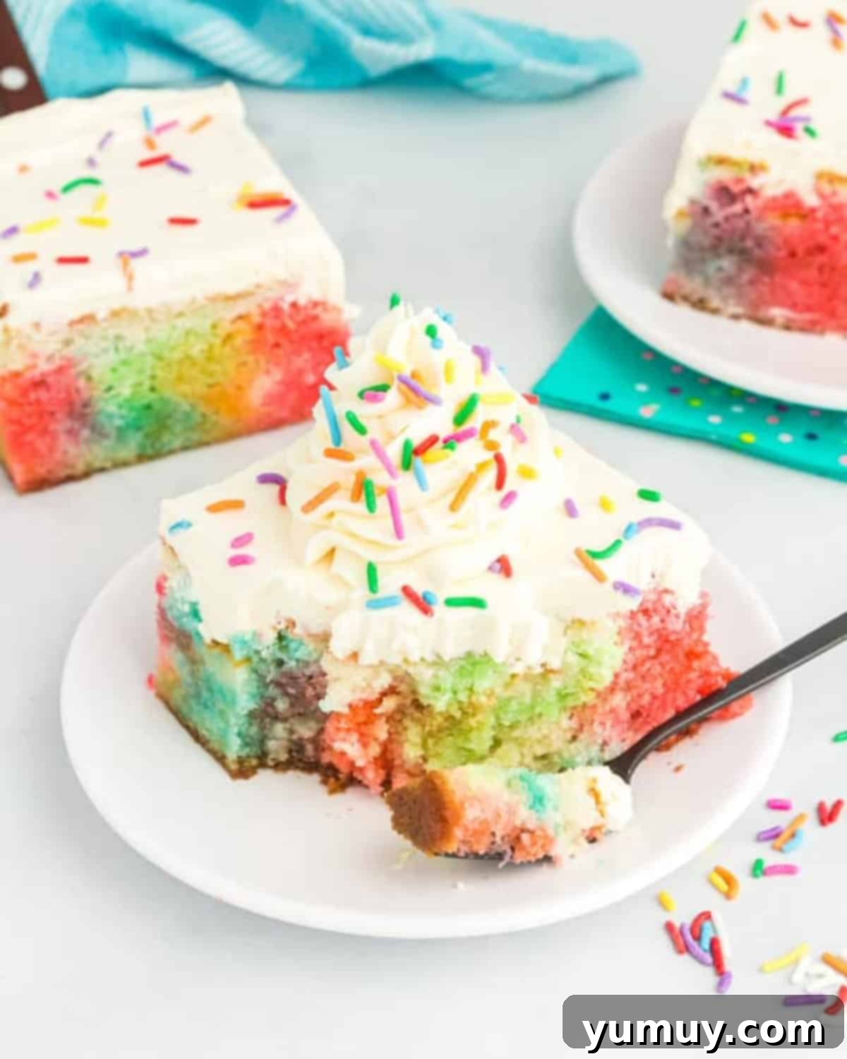 A close-up view of a slice of rainbow poke cake on a plate with a bite taken out, showcasing the distinct layers of colored Jell-O within the cake and the smooth frosting.