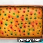 An overhead view of a vanilla cake in a rectangular pan with holes being filled by colorful Jell-O liquids, creating a rainbow pattern.