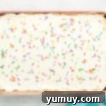 An overhead shot of the fully frosted rainbow poke cake in a rectangular baking pan, adorned with colorful sprinkles, ready to be sliced.