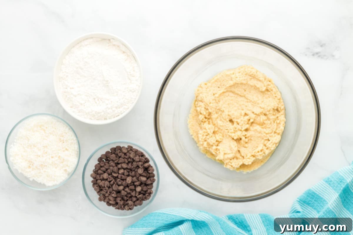 Tropical Coconut Chocolate Chip Delight 13 dry ingredients added to coconut chocolate chip cookie dough in a glass bowl, partially mixed.