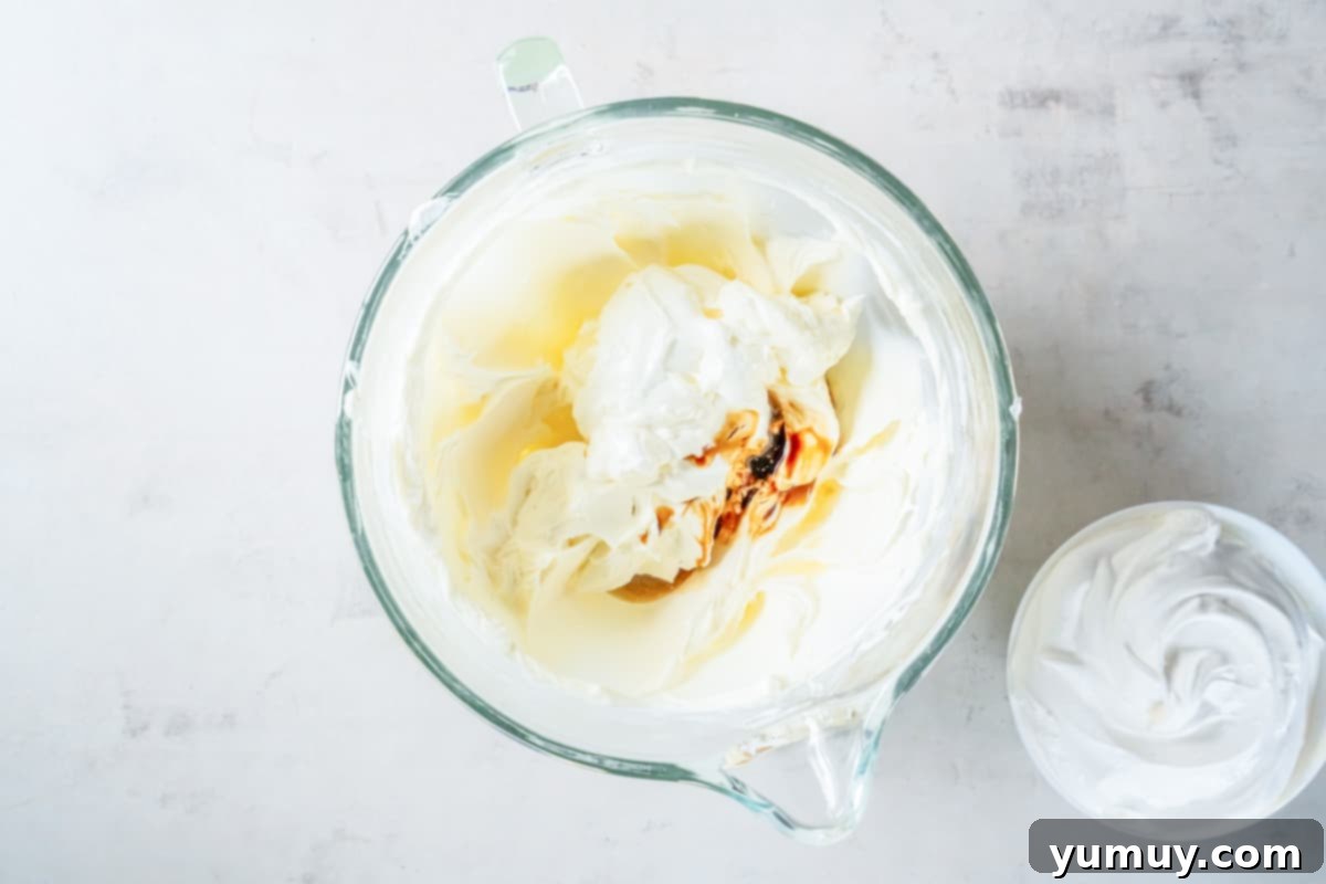 Fresh lemon juice, pure vanilla extract, and creamy sour cream being poured into a glass bowl containing the whipped cream cheese mixture, adding essential flavors.