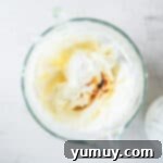Lemon juice, vanilla extract, and sour cream being added to the creamy cream cheese mixture in a clear glass bowl, ready for the next step.