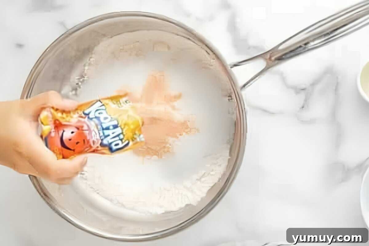 Fun Fragrant Kool Aid Playdough 11 Adding a packet of vibrant Kool-Aid powder to a saucepan containing flour and other dry ingredients for playdough.