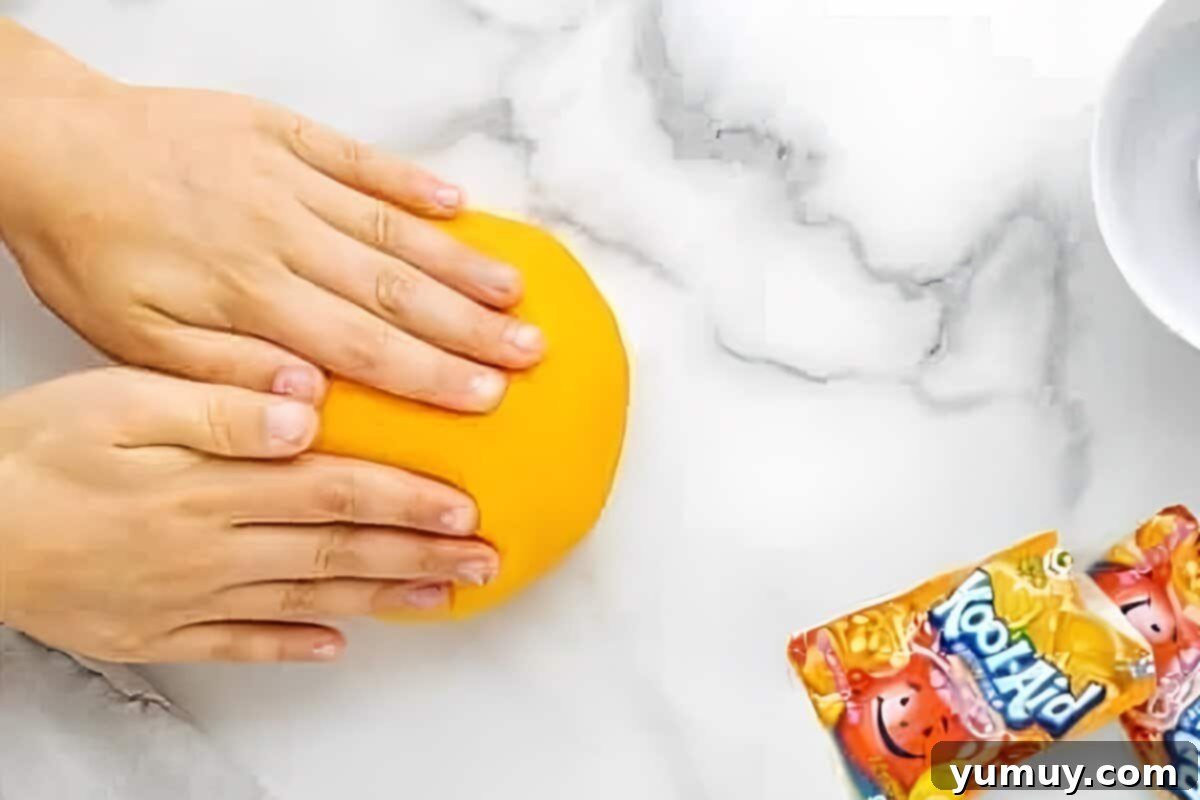 Fun Fragrant Kool Aid Playdough 14 Hands expertly kneading a warm, vibrant orange ball of homemade playdough on parchment paper until it reaches a smooth, pliable consistency.