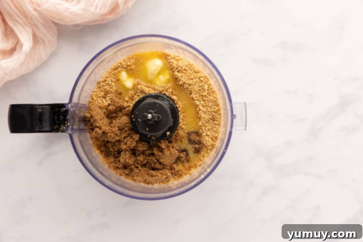 Graham cracker crumbs, brown sugar, and melted butter being processed in a food processor, resembling damp sand.