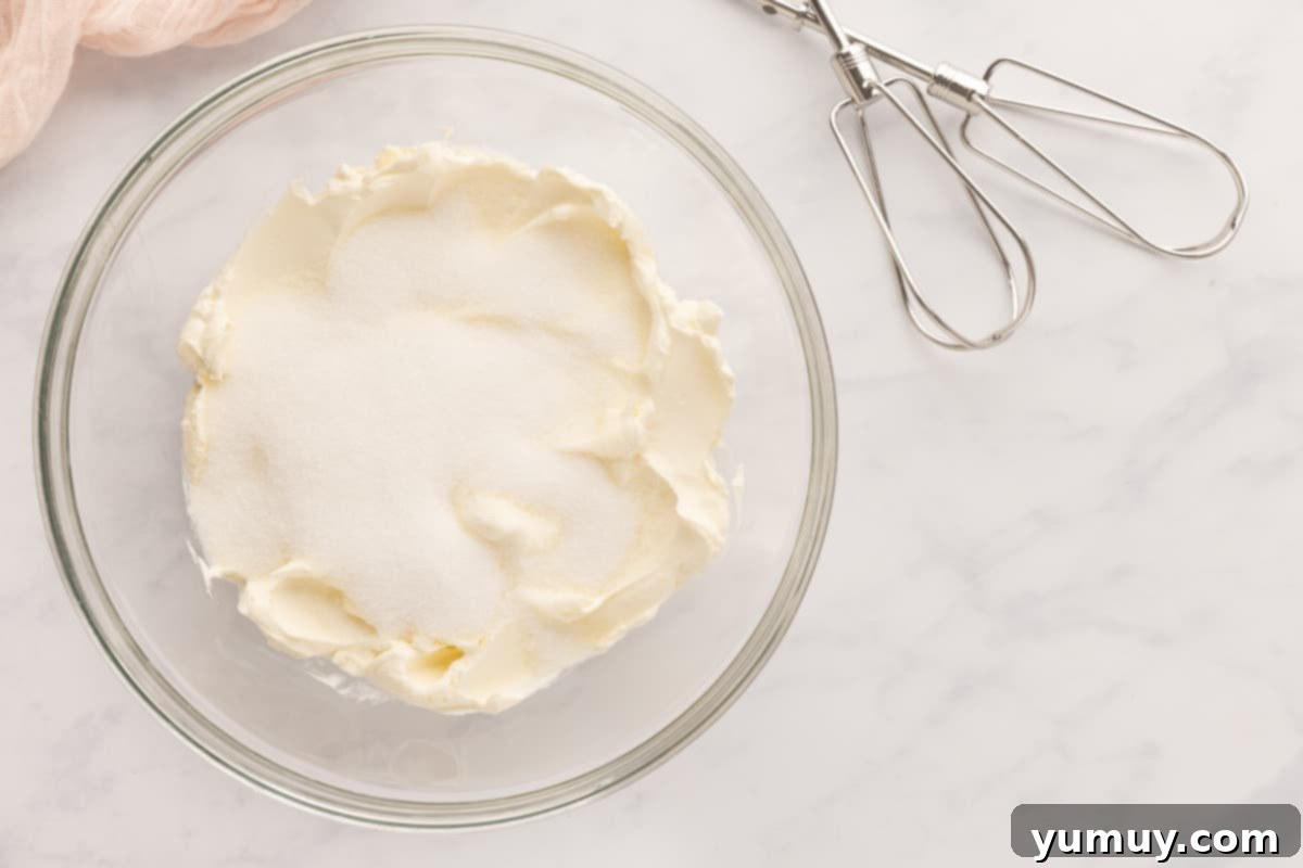 Smooth, creamy whipped cream cheese and sugar in a glass bowl, with a whisk attachment from a hand mixer resting nearby.