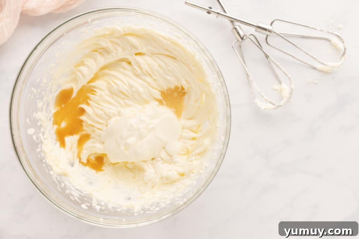 A glass bowl containing a light, creamy mixture of cream cheese, sour cream, vanilla, and lemon juice, partially blended with a hand mixer.