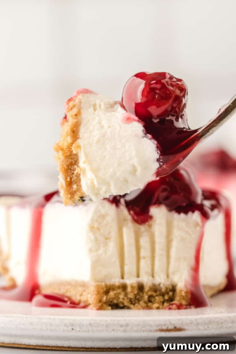 A pristine slice of no-bake cherry cheesecake, adorned with a glossy cherry topping, rests on a white plate next to a fork, ready to be enjoyed.