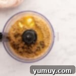 A food processor bowl containing graham cracker crumbs, brown sugar, and melted butter mixed to a damp sand consistency.
