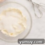 Cream cheese and sugar being beaten in a glass bowl with a hand mixer, creating a smooth, creamy texture.