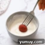 A red velvet cake truffle being dipped into melted white chocolate using a fork, ensuring full coverage and a smooth finish.