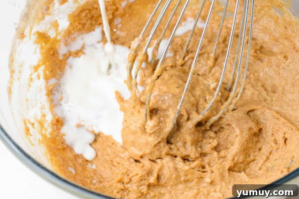Milk being added to a mixed batter in a glass bowl, stirred with a whisk.