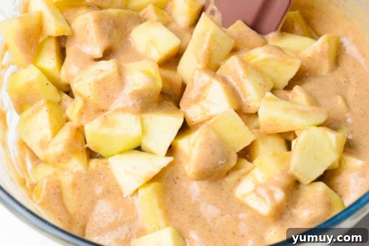 Chopped apple pieces being gently folded into the fritter batter in a glass bowl with a spatula.