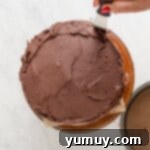 A hand expertly spreading chocolate frosting onto a chocolate cake with an offset spatula to achieve a smooth finish.