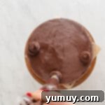 A hand artistically piping chocolate frosting decorations onto a chocolate cookie dough layer cake on a wooden cake stand.