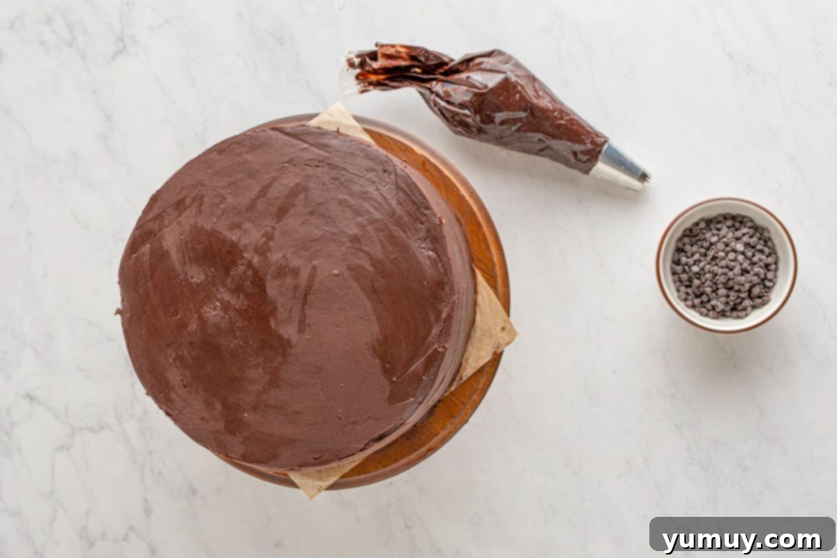 A chocolate cookie dough layer cake positioned on a wooden cake stand, partially covered with a thin crumb coat of chocolate frosting, with a piping bag filled with more frosting nearby.