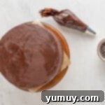 A chocolate cookie dough layer cake on a wooden cake stand, partially covered with a crumb coat of chocolate frosting, with a piping bag ready for decoration.