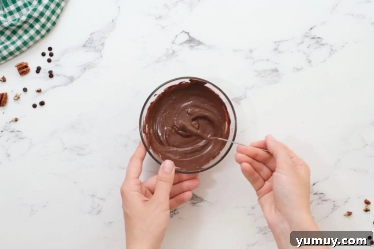 Irresistible Homemade Turtle Clusters 16 A spoon stirring rich, melted chocolate in a clear glass bowl, showcasing its smooth consistency.