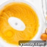 Whole eggs being carefully added, one by one, to the vibrant orange cheesecake filling in a mixing bowl.